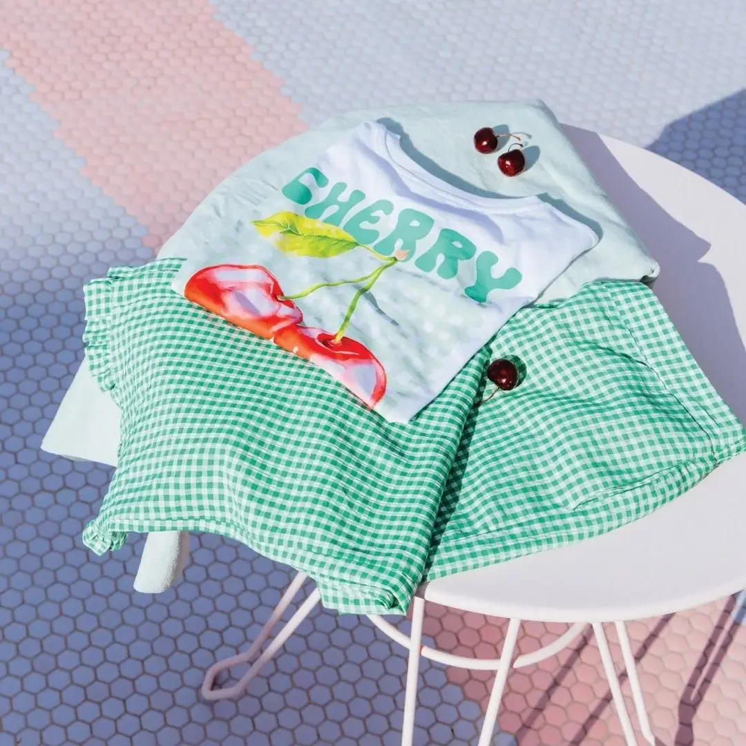 Flatlay d’un look estival Cache Cache avec t-shirt cerises et jupe vichy verte